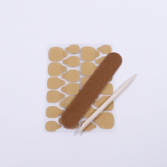 Un kit press on nails complet présenté à plat sur fond blanc : lime marron, gels pads et bâtonnet de buis, tout le nécessaire pour une pose de faux ongles réussie à domicile.